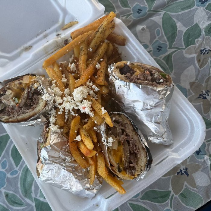 A wrap cut into three pieces in a takeout container, each piece still half in foil and showing chopped steak, melted cheese, onion and peppers, with seasoned french fries piled in the middle under crumbled feta