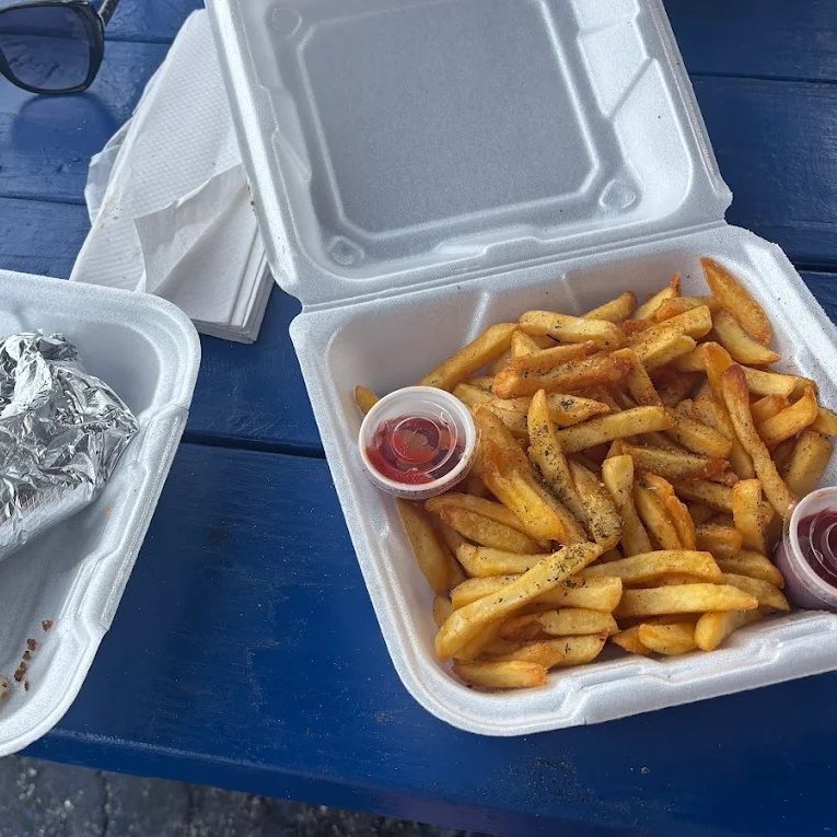 Two open takeout containers on a blue picnic table — a foil-wrapped gyro in one, a large portion of seasoned Greek fries with two cups of ketchup in the other