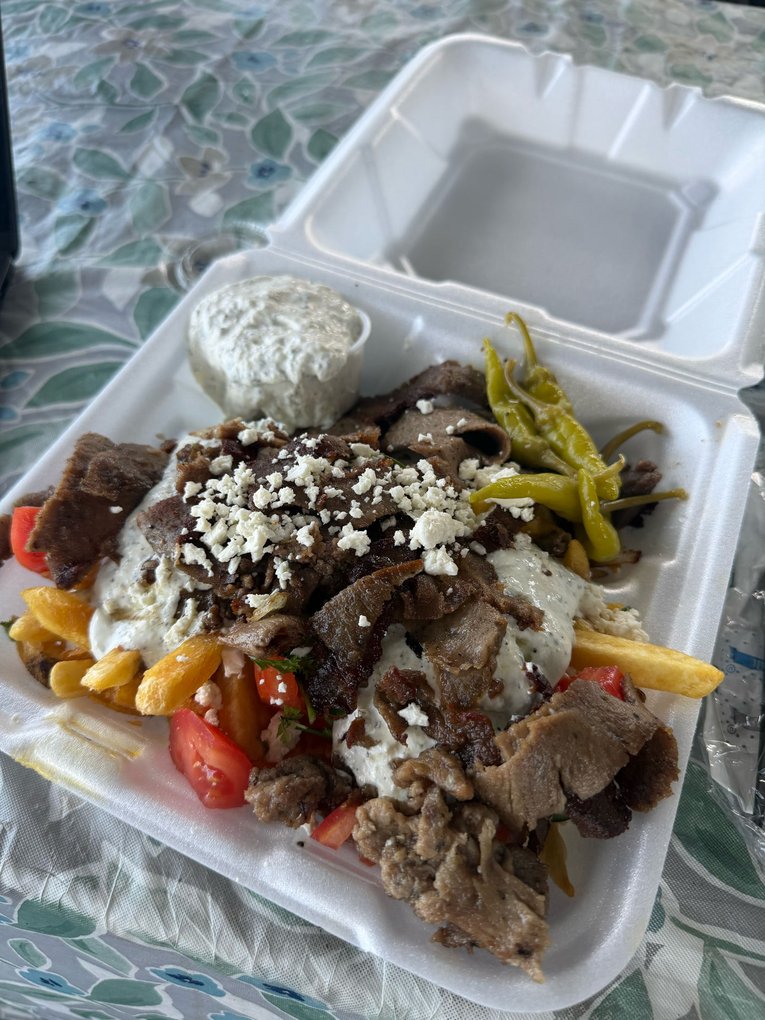 An open takeout container piled with sliced lamb and chicken gyro meat, a scoop of tzatziki, crumbled feta, three pepperoncini peppers and tomato wedges over french fries, with a second cup of tzatziki beside it