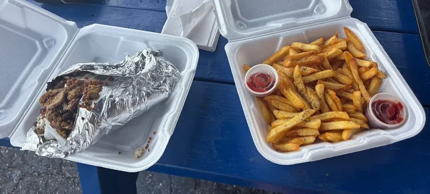 A foil-wrapped gyro in an open takeaway box beside a box of seasoned Greek fries with two cups of ketchup, on the truck's blue picnic table
